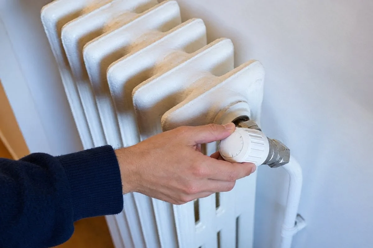 Mano que ajusta la válvula termostática de un radiador blanco de hierro fundido, junto a la pared